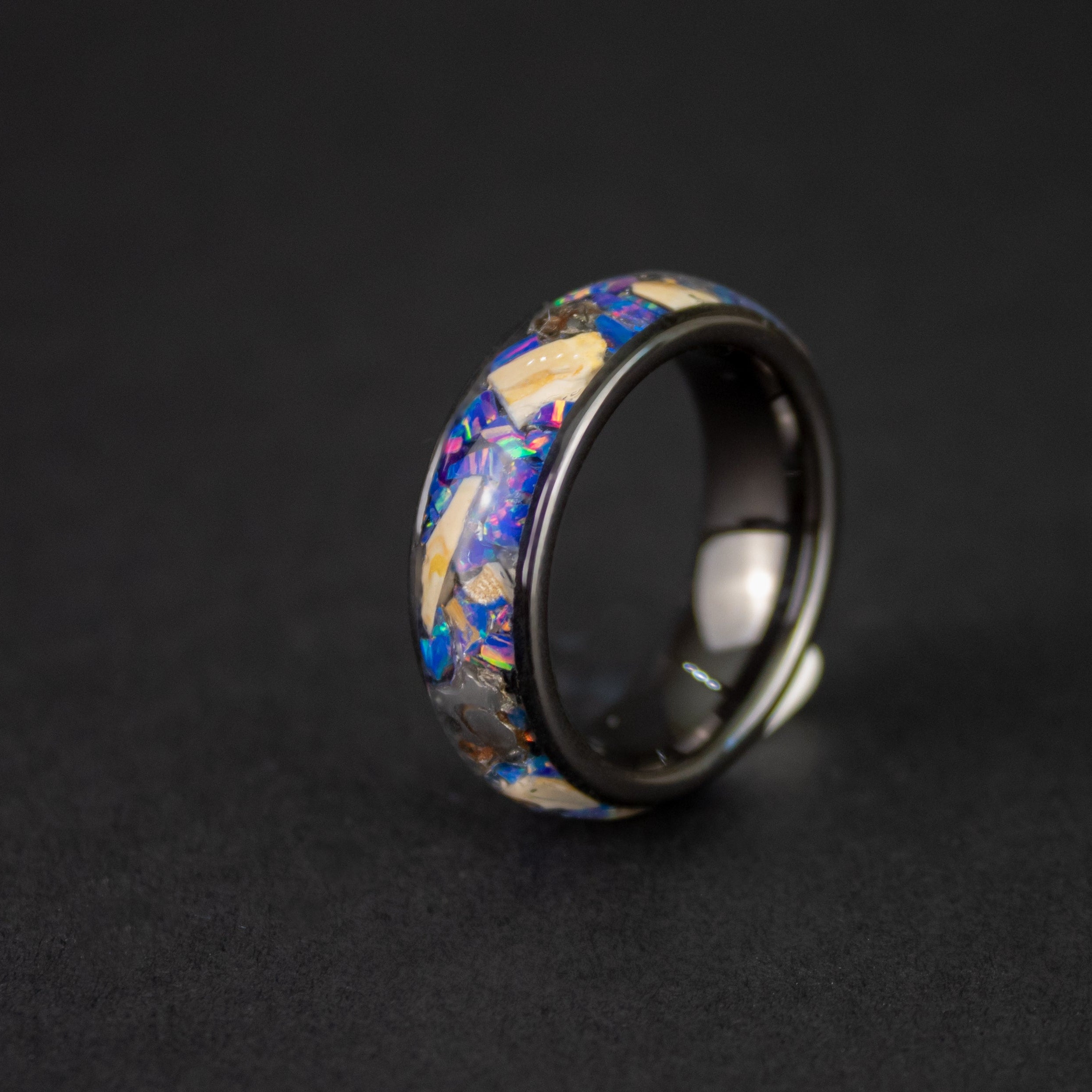 Detail shot of the Midnight Mosasaur ring showing large authentic Mosasaur bone pieces and meteorite chunks catching light through midnight blue purple opal inlay on wide channel black ceramic band