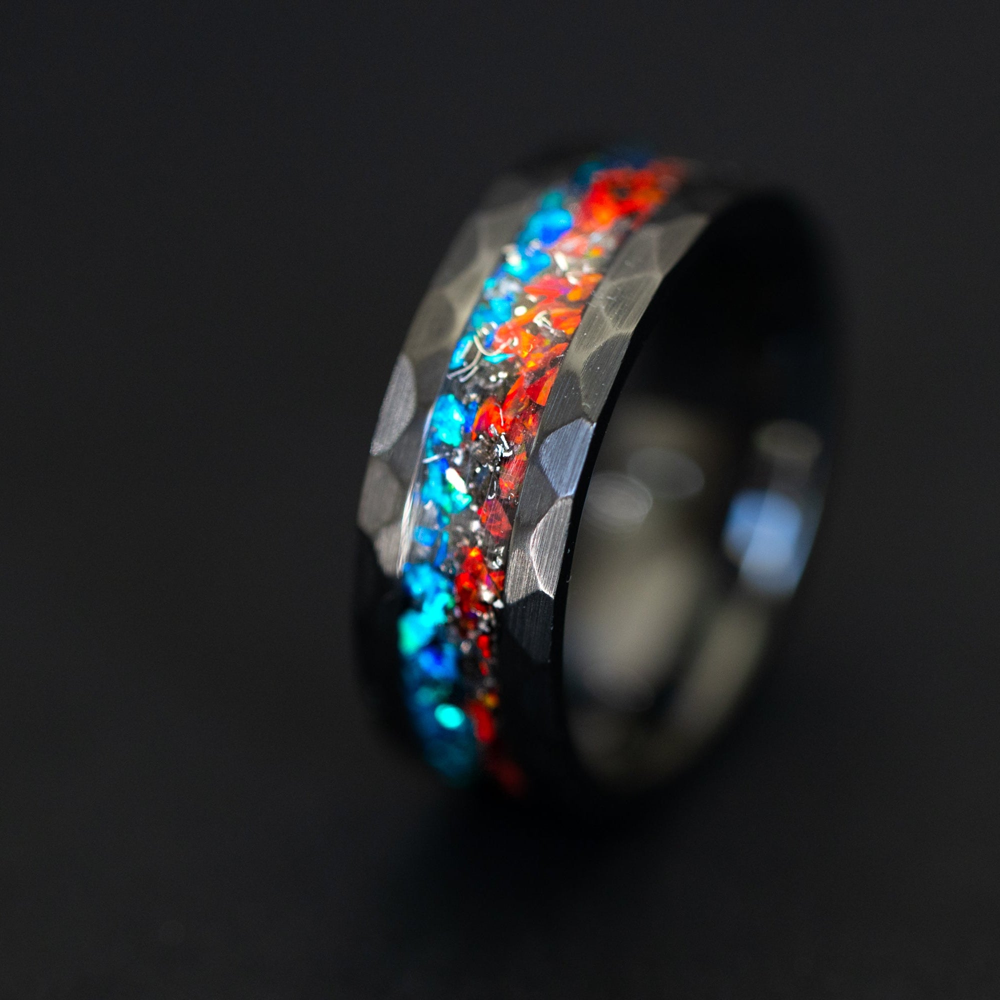 Close up of meteorite shavings and contrasting red and blue opals in a hammered black ceramic ring.